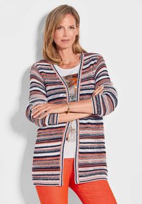 Gestreepte cardigan in marineblauw, oranje en wit; open voorkant, geribde manchetten, structuurgebreid; gedragen over een witte top en oranje broek.