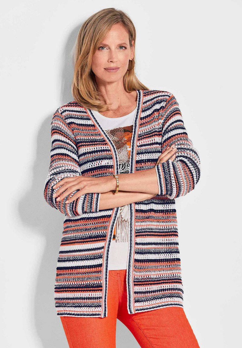 Gestreepte cardigan in marineblauw, oranje en wit; open voorkant, geribde manchetten, structuurgebreid; gedragen over een witte top en oranje broek.