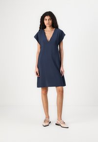Rochie scurtă de culoare bleumarin cu decolteu în V, mâneci ample și tiv ușor evazat. Material moale cu o textură netedă. Asortată cu încălțări de o nuanță deschisă.