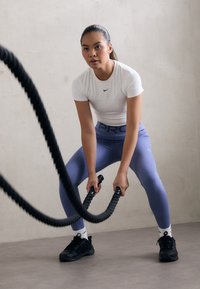 Žena u bijelom Nike majici i plavim tajicama drži crne bojne užadi. U pozadini je svijetla teksturirana zidna površina i sivi pod.