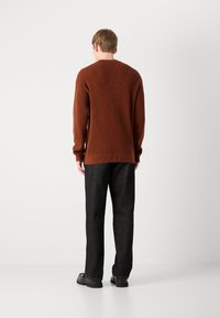 Bruine gebreide trui met een geribbelde textuur, gecombineerd met donkere, losse broek, gezien van achteren. Zwarte schoenen zichtbaar.