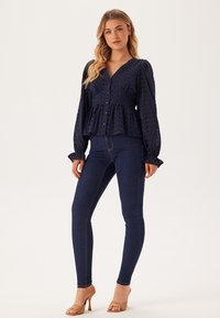 Marineblauwe blouse met knopen, afgewerkt met gestructureerde stippen, peplum taille en pofmouwen, gecombineerd met donkere skinny jeans en nude hakken.