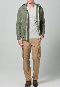 Olijfgroene jas met capuchon, ritssluitingen en voorzakken, gecombineerd met een lichtgrijs T-shirt en beige cargobroek. Bruine schoenen maken de outfit compleet.