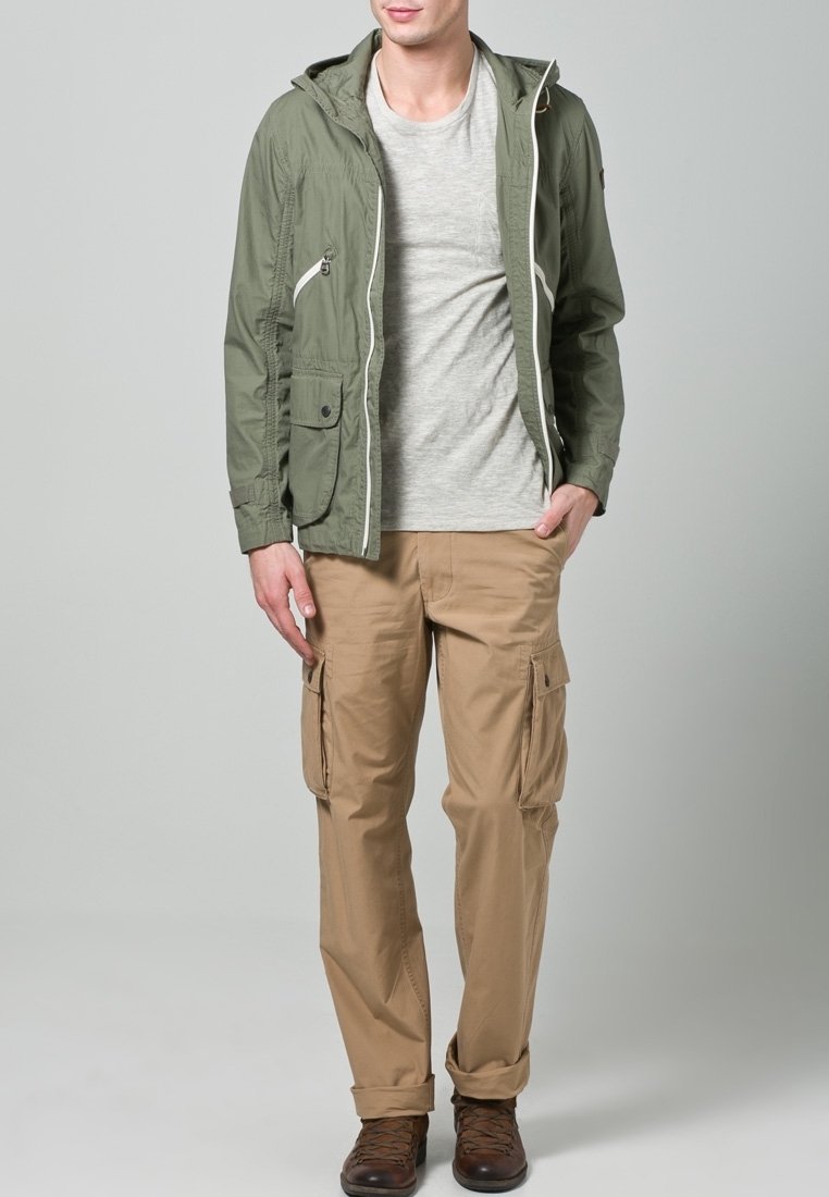 Olijfgroene jas met capuchon, ritssluitingen en voorzakken, gecombineerd met een lichtgrijs T-shirt en beige cargobroek. Bruine schoenen maken de outfit compleet.