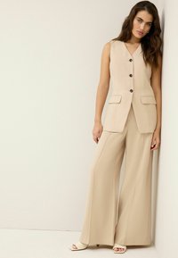 Gilet beige à rayures avec un revers cranté, boutons noirs et deux poches avant, associé à un pantalon large assorti et des sandales légères.