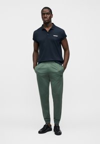 Μπλε σκούρο πόλο μπλούζα με κοντά ρυμουλκούμενα μανίκια, πράσινα στενά αθλητικά παντελόνια και μαύρα αθλητικά παπούτσια. Διαθέτει μικρό λογότυπο στη μπλούζα.