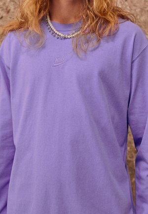 Persoon in een paarse Nike longsleeve shirt en gelaagde kettingen met parels en zilveren spikes, tegen een vervaagde achtergrond.