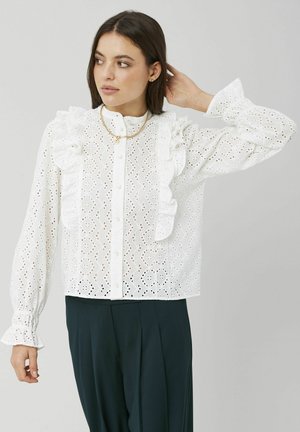 Blouse blanche avec bordure à volants, motif floral en broderie anglaise, boutonnière à l'avant et poignets élastiques. Portée avec un pantalon vert foncé et un collier superposé.