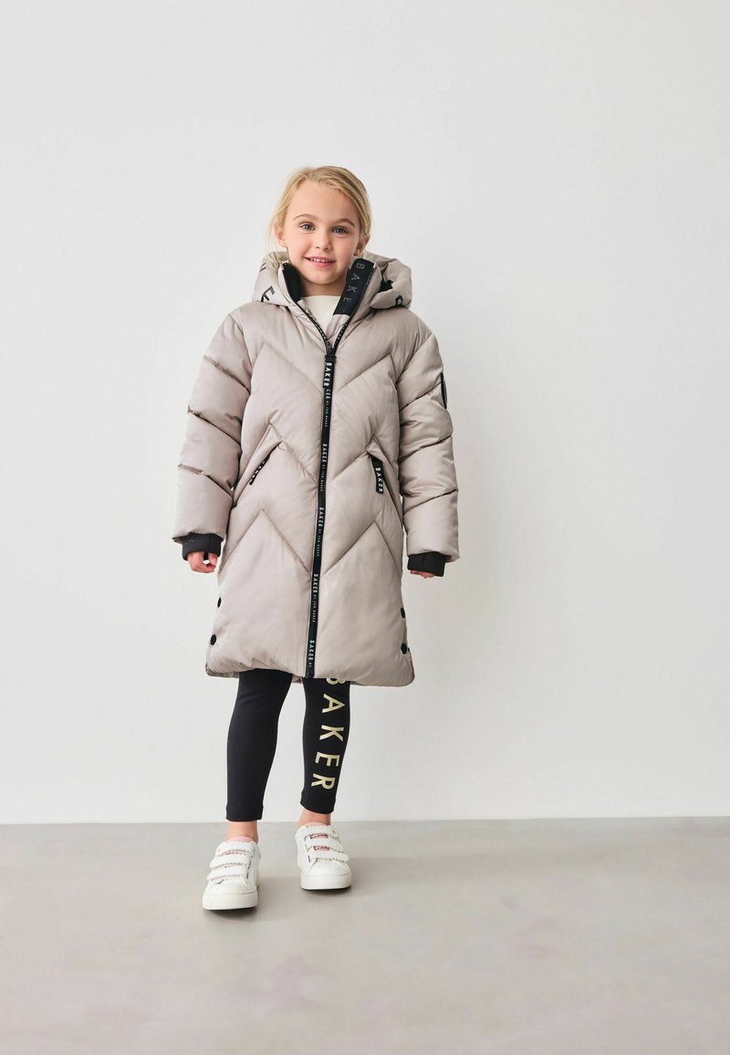 Cappotto beige imbottito con trapuntatura diagonale, zip nera e cappuccio, abbinato a leggings neri con testo dorato e scarpe bianche.