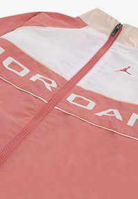 Gaišs, kabatā ar rāvējslēdzēju, jakas korallkrāsā un baltā krāsā, ar augstu apkakli, lielu "JORDAN" logotipu un mazu Jumpman logotipu sarkanā krāsā. Gluda tekstūra.