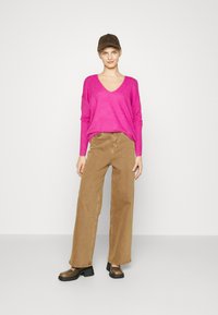 Fuchsia V-hals trui, losse pasvorm, gecombineerd met tan brede broek en stevige zwarte hakken. Model draagt een bruine pet en staat tegen een effen achtergrond.