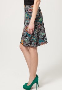Jupe noire évasée avec des motifs floraux colorés en vert, orange et bleu. Caractérisée par un ourlet festonné et associée à des escarpins verts.