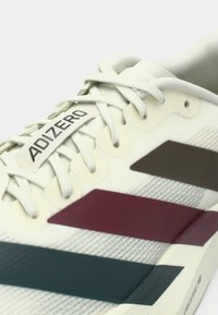 Κοντινό πλάνο άσπρου Adidas Adizero παπουτσιού με μπορντώ και μαύρες λωρίδες και άσπρα κορδόνια σε ανοιχτό φόντο.
