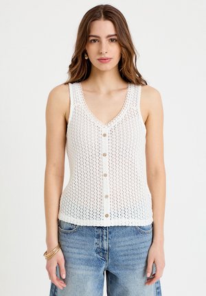 Kobieta w białym, bezrękawnym, szydełkowanym topie na guziki i niebieskich dżinsach, stojąca na jednolitym tle.