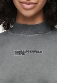 Sivá mikina s vysokým rebrovaným golierom. Na hrudi je vyšitý nápis "KARL LAGERFELD JEANS" v čiernej farbe. Mäkka textúra látky.