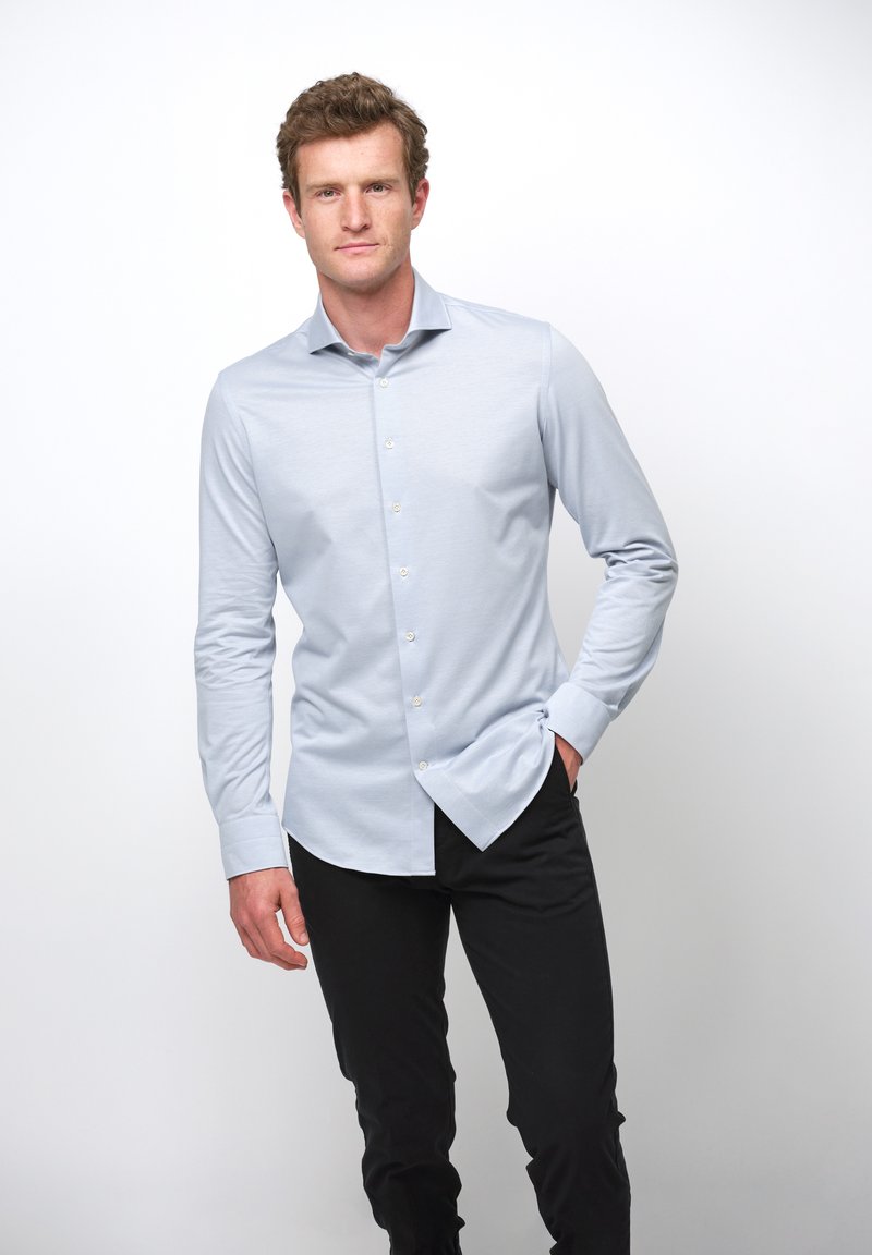 Camisa de manga larga azul claro con botones en la parte delantera y cuello puntiagudo, hecha de una tela suave. Combinada con pantalones negros, el modelo está de pie de manera relajada con una mano en el bolsillo.