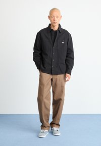 Black button-up shirt with two front pockets, paired with brown pants. The outfit features gray shoes with white soles on a blue floor.
