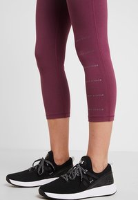 Bordowe legginsy capri z subtelnym logo marki po bokach, zestawione z czarnymi butami sportowymi z białymi podeszwami i wzorzystymi sznurowadłami.