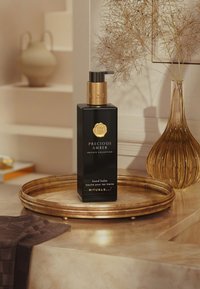 Svart rektangulär flaska med handbalm med en guldpump och präglad logotyp; placerad på ett gyllene fat med en texturerad brun duk i närheten.