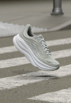 Gaiši pelēka skriešanas apavu ar bieza baltas zolītes, kurā rakstīts "HOKA", virs gājēju pārejas uz asfalta.