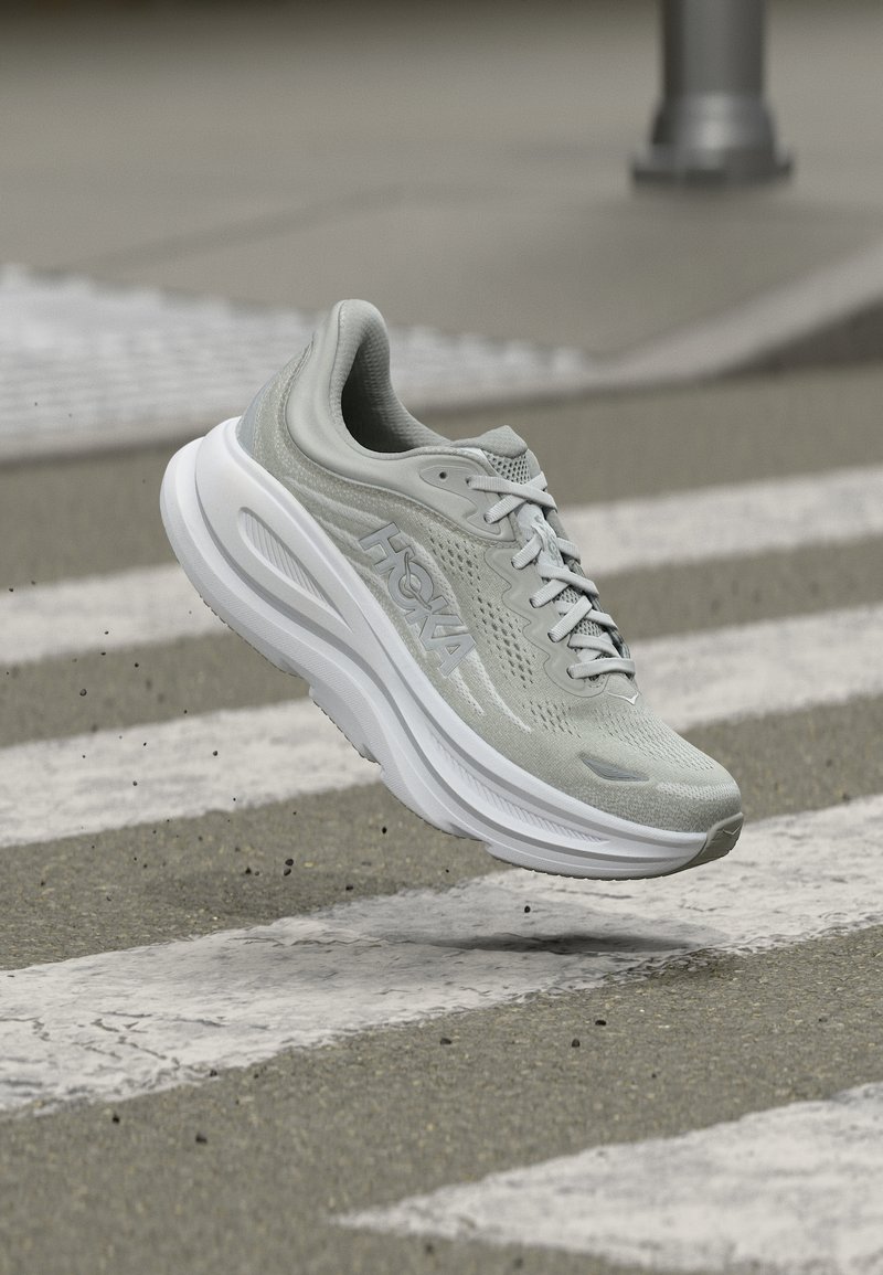 Ανοιχτό γκρι παπούτσι για τρέξιμο με παχιά λευκή σόλα, επώνυμο "HOKA", αιωρείται πάνω από διάβαση πεζών σε άσφαλτο.