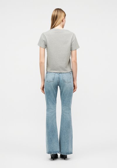 Γυναίκα που στέκεται με την πλάτη, φορά γκρι κοντομάνικο μπλουζάκι, ανοιχτό μπλε τζιν καμπάνες και μαύρες μυτερές γόβες επάνω σε λευκό φόντο.