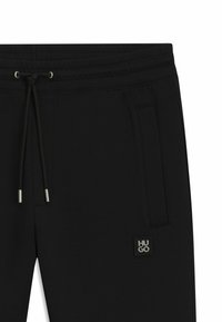 Black jogger pants made of soft fabric, featuring an elastic waistband with drawstring, side pockets, and a branded logo patch.