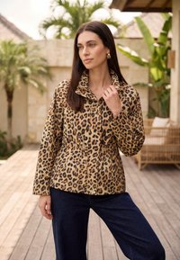 Femme aux cheveux bruns lisses portant de grandes boucles d'oreilles en or et une veste à imprimé léopard, debout sur un patio en bois entouré de plantes tropicales.