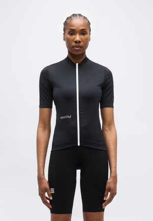 Frau steht mit Blick nach vorne, trägt ein schwarzes Radtrikot mit kurzen Ärmeln und durchgehendem Frontreißverschluss sowie passende schwarze Radhosen.