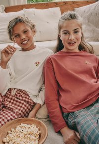 Zwei Mädchen sitzen zusammen, tragen ein weißes Langarmshirt mit floralen Stickereien und ein rostfarbenes Shirt, dazu karierte Schlafanzughosen.