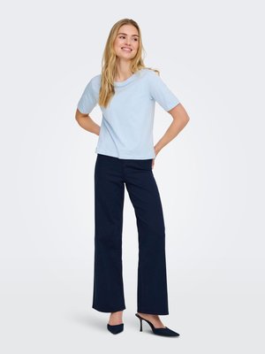 Young woman smiling with long blonde hair wearing a light blue short-sleeve shirt, navy wide-leg pants, and black high-heeled shoes.