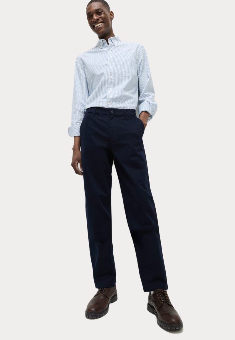 Camicia azzurra a righe con bottoni, pantaloni blu scuro e stivaletti marroni con lacci. L'outfit presenta una vestibilità comoda e uno stile casual.