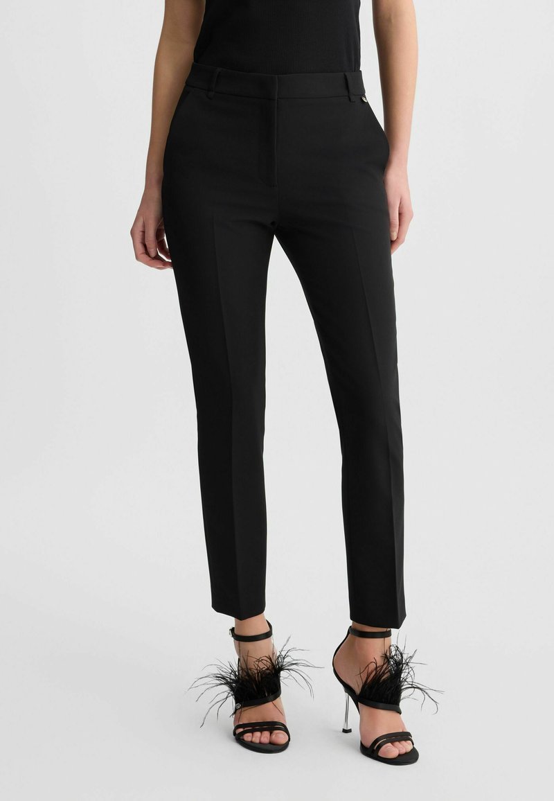 Zwarte op maat gemaakte broek met een slim fit, gemaakt van soepel materiaal en voorzien van voorplooien. Gecombineerd met zwarte sandalen met hakken, versierd met veeraccenten.