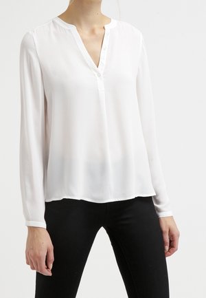 Femme portant un chemisier blanc à manches longues avec col en V et ouverture partielle à boutons à l'avant, associé à un pantalon noir.