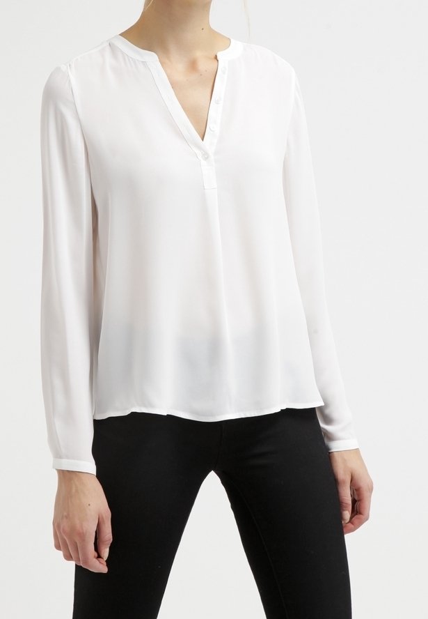 Femme portant un chemisier blanc à manches longues avec col en V et ouverture partielle à boutons à l'avant, associé à un pantalon noir.