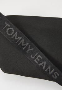 Schwarze Nylon-Tasche mit texturierter Oberfläche, die ein auffälliges graues "TOMMY JEANS"-Logo am Tragegurt trägt. Rechteckige Form mit minimalistischem Design.