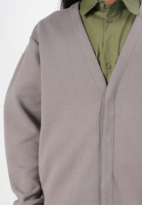 Ljusgrå cardigan i mjukt tyg, med V-krage, långa ärmar och avslappnad passform. Bärs över en grön skjorta med krage.