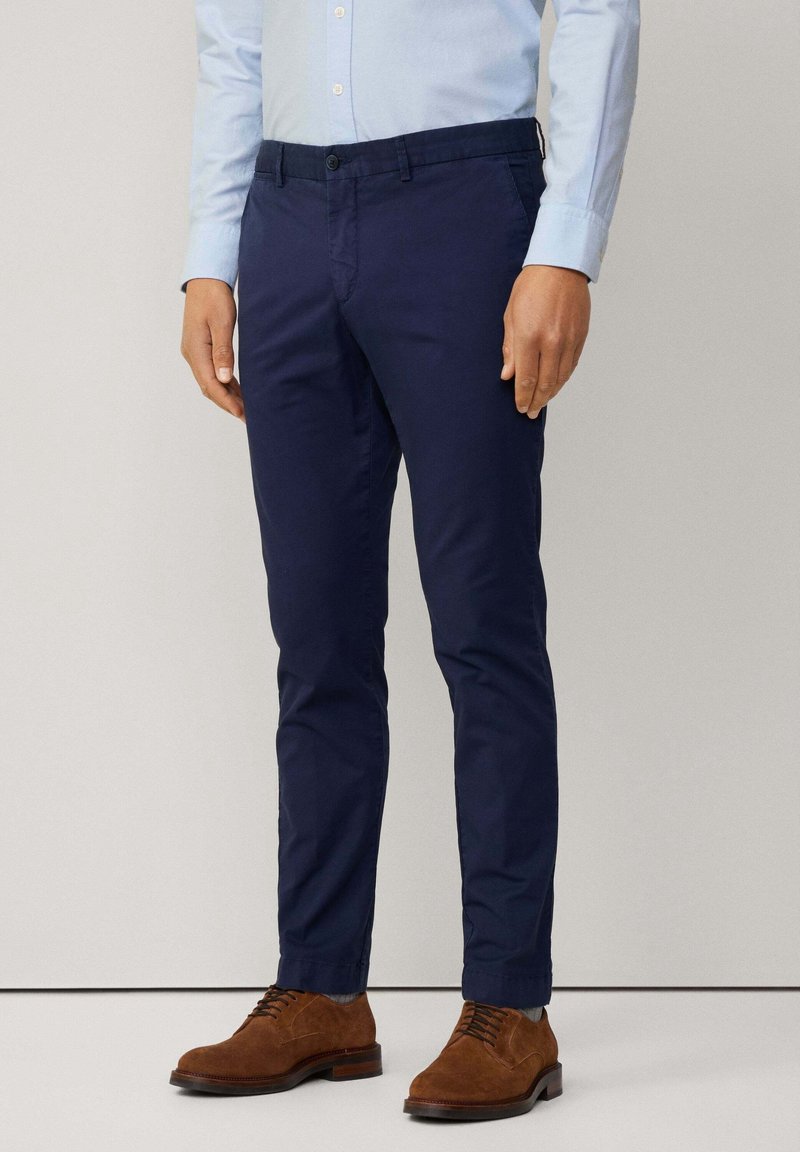 Homme portant un pantalon slim bleu foncé, une chemise bleu clair boutonnée et des chaussures habillées en daim marron, debout devant un fond uni.