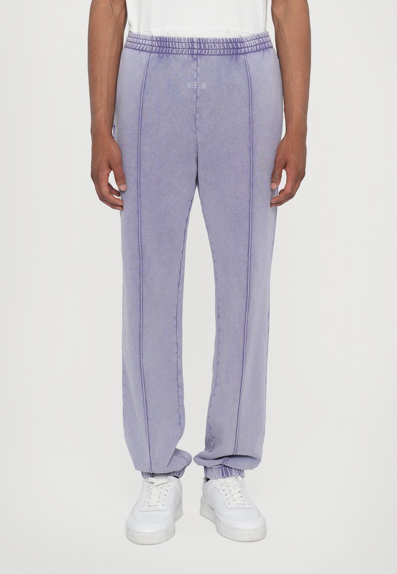 Light purple sweatpants feature an elastic waistband, white stitching details, and cuffs at the ankles, paired with white sneakers.