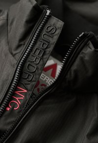 Chaqueta negra con cremallera, de tejido texturizado, que cuenta con un alto cuello y bordado de marca en blanco y rojo a lo largo de la cremallera.