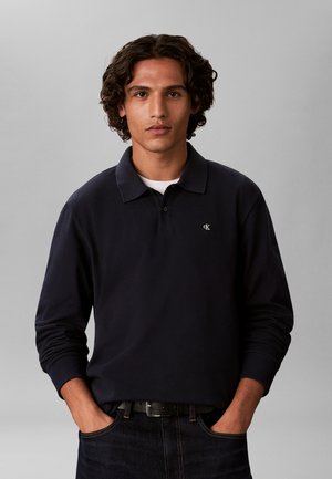 Jeune homme aux cheveux bouclés portant un polo noir à manches longues avec un t-shirt blanc en dessous, mains dans les poches, fond gris neutre.