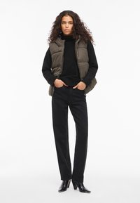 Prsluk u maslinasto zelenoj boji preko crnog džempera, uparen s crnim širokim hlačama i crnim čizmama s potpeticom. Glatke teksture, minimalni detalji.