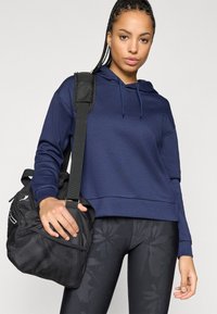 Cropped hoodie azul-marinho com capuz ajustável, combinado com uma mala de duffel preta. Leggings pretas com um padrão floral subtil.