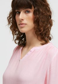 Frau mit lockigem braunem Haar, die eine hellrosa Bluse mit kleinem V-Ausschnitt trägt und leicht zur Seite blickt, vor einem einfarbigen Hintergrund.