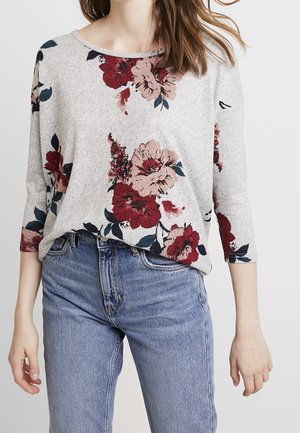 Vrouw draagt een lichtgrijs shirt met lange mouwen, bedrukt met rode en roze bloemen, gecombineerd met een hoge taille blauwe spijkerbroek.
