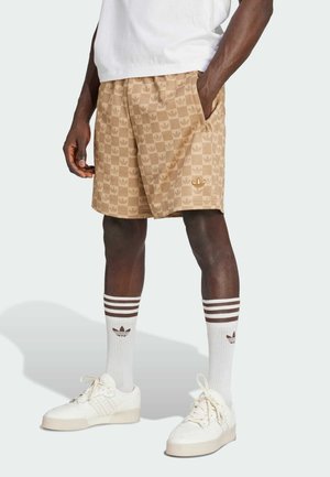 Mężczyzna ubrany w beżowe kratkowane szorty z logo Adidas, białe skarpetki Adidas z brązowymi paskami oraz kremowe buty sportowe Adidas, stoi z rękami w kieszeniach.