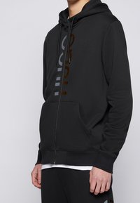 Felpa nera con zip in tessuto morbido, con tasca frontale e stampe del logo a contrasto in grigio e marrone sul lato sinistro.