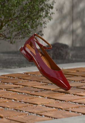 Ballerine plate rouge brillante avec bride à la cheville flottant au-dessus d'un bloc de bois et d'une surface extérieure en galets, arbuste vert flou et pierre en arrière-plan.