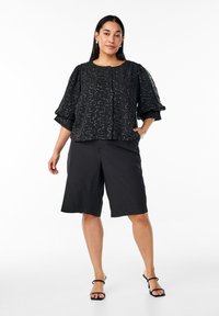 Sort knap-op skjorte med tekstureret stof og puffede ærmer, parret med knælange sorte shorts og stribede sorte sandaler.