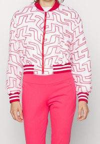 Bílá bomber bunda s červeným zigzag vzorem, žebrované červenobílé pruhované manžety, zipové zapínání a nabírané rukávy. Nosit s růžovými kalhotami.